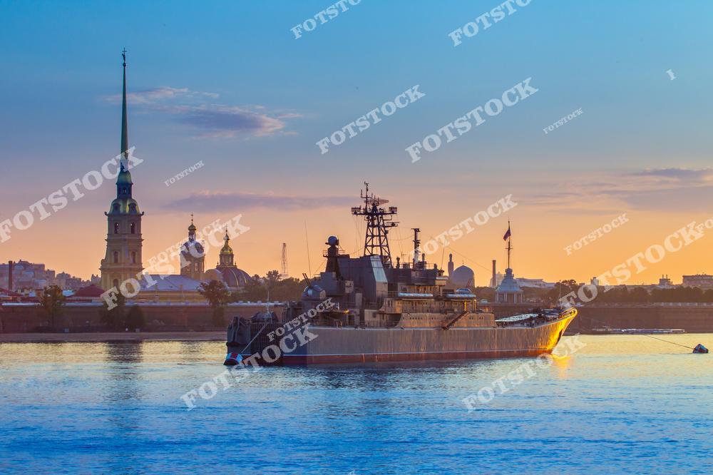 На фото изображен военный корабль, стоящий на Неве на фоне Петропавловской крепости. Высокий шпиль Петропавловского собора ярко выделяется в закатных лучах, отражаясь в спокойной воде реки. Вечернее небо придаёт кадру теплые тона, создавая контраст с величественными очертаниями крепости и военного судна. Этот вид передает уникальную атмосферу Санкт-Петербурга, сочетающую силу и историю.